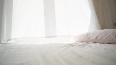 Güneşli bir sabah, pencereden gelen ışık beyaz yatağa düşer, rüzgar perdeleri savurur. Kamera yatağın üstünde çekim yapar, yakın plan.