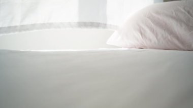 Güneşli bir sabah, pencereden gelen ışık beyaz yatağa düşer, rüzgar perdeleri savurur. Kamera yatağın üstünde çekim yapar, yakın plan.