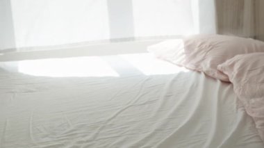 Güneşli bir sabah, pencereden gelen ışık beyaz yatağa düşer, rüzgar perdeleri savurur. Yataktan pencereye genel bakış.