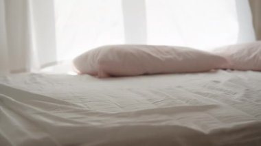 Güneşli bir sabah, pencereden gelen ışık beyaz yatağa düşer, rüzgar perdeleri savurur. Yatakta pembe bir yastık alıyorum, ağır çekimde..