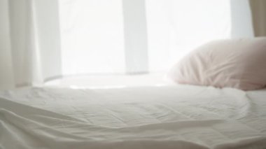 Güneşli bir sabah, pencereden gelen ışık beyaz yatağa düşer, rüzgar perdeleri uçurur, yakın plan..