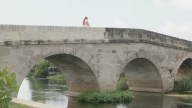 Nehir boyunca uzanan üç kemerli tuğla bir köprü. Genç kadın üzerinde yürüyor. Yaz, sudaki çimenler. Yavaş çekim
