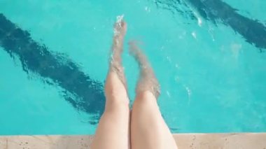 Genç kadın havuzun kenarında oturuyor ve ayaklarını suda duruluyor. Yukarıdan yakın çekim, yavaş çekim. Güneşli yaz.