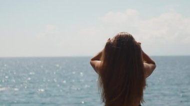 Deniz kenarında mayo giymiş tombul genç bir kadın topuz şeklinde uzun saçlar topluyor..