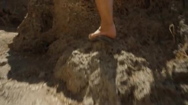 Genç kadın deniz kenarındaki kayalıklara tırmanıyor, kamera ayaklarını takip ediyor..