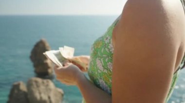 Denizde uçurumun kenarında genç bir kadın para sayıyor, kamera daireler çiziyor..