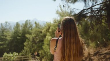 Çam ormanındaki genç bir kadın dağları telefonla izliyor. Güneşli hava.