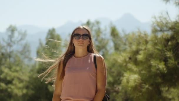 一个年轻的女人在松树林中背靠着高山 她的头发在风中飘扬 她把它们理直了 — 图库视频影像