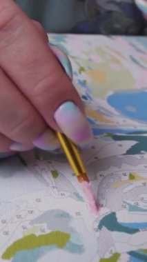 Genç kadın bir resmi fırçayla boyayla numaralarla boyuyor, hareket halinde, yakın plan. Dikey video.