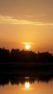 Zaman ayarlı. Gölde gün doğumu, ağaçlardan yükselen güneş bulutlara karışıyor. Dikey video.
