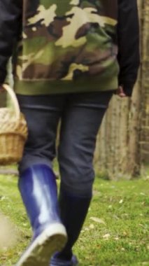 Kamuflaj ceketli genç bir kadın elinde sepetle ormanda yürüyor. Mantar arıyorum. Dikey video.