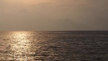 Denizde altın gün batımı, bulutların arasından güneş doğuyor. Arkaplanda dağlar.