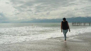 Tombul genç bir kadın, dağların ve gök gürültüsü bulutlarının arkaplanında ağır çekimde deniz kıyısında yürüyor..