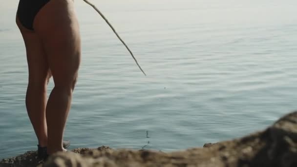 一个穿着泳衣的年轻女子正在悬崖边钓鱼 拿着钓竿 看着浮子 — 图库视频影像
