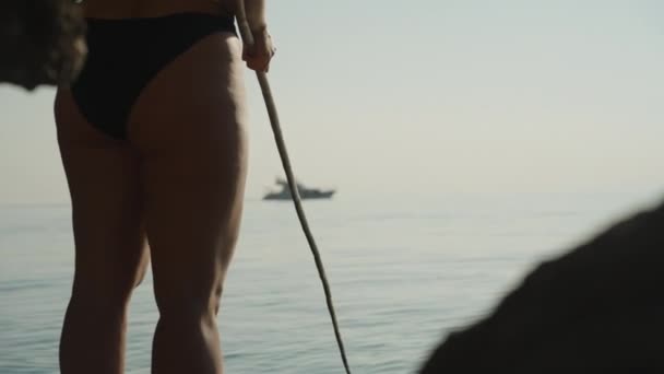 一个穿着泳衣的年轻女子正在悬崖边钓鱼 她拿出一根钓竿 一艘游艇在船尾航行 — 图库视频影像