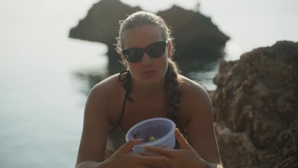一个穿着泳衣的年轻女子正坐在海里的岩石上 后面的水里有一块岩石 她递给我一盘葡萄 — 图库视频影像