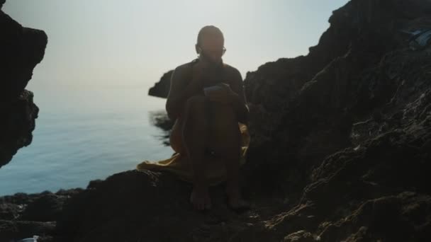 一个穿着泳衣的年轻女子正坐在海中的岩石上吃葡萄 阳光在她身后 — 图库视频影像
