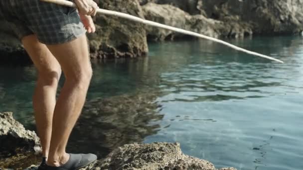 ショートパンツの男は自家製の竹釣り棒で海で釣りをしている 彼は水で石の上に座る — ストック動画
