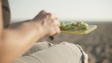 Denizde güneşli bir hava, genç bir kadın sahilde salata yapıyor. Dilimlenmiş avokadoyu salata kasesine koyar..