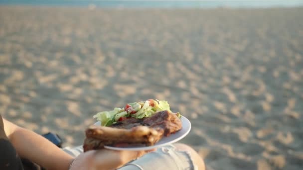 海でビーチでピクニック 若い女性はステーキとサラダの皿を持っています クローズアップ — ストック動画