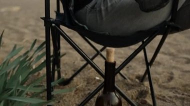 Bir kız sahilde bir sandalyede oturuyor ve şarap içiyor. Kumsalda bir şişe şarap.