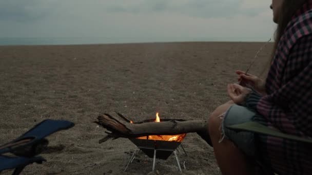 トワイライト 海でビーチでピクニック 火を見ている椅子の若い女性と彼女の手で棒を回す — ストック動画