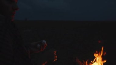 Deniz kenarında gece pikniği. Ekoseli genç bir kadın çubuğa marşmelov ekiyor. Ateş