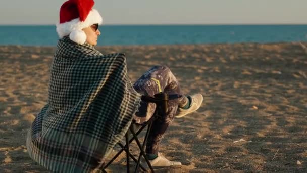 サンタの帽子をかぶった孤独な若い女性は 海でビーチで新年を祝います 毛布に包まれる — ストック動画