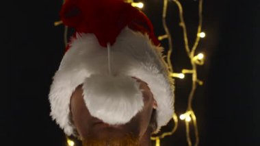 Kırmızı sakallı bir adam Noel Baba şapkası takıyor ve burnunda ponpon var, gülümsüyor. Siyah arka plan ve yanıp sönen ışıklar. Yakından, korkutucu..