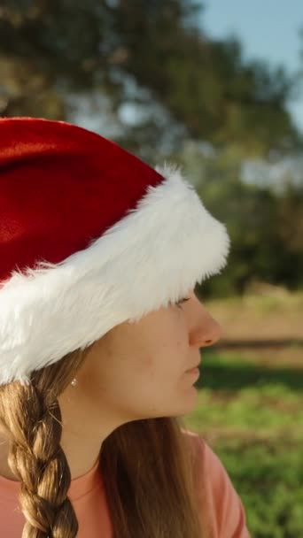 バーティカルビデオ 赤いサンタの帽子をかぶった若い女性に髪を編みました クリスマスの森でヨガ — ストック動画