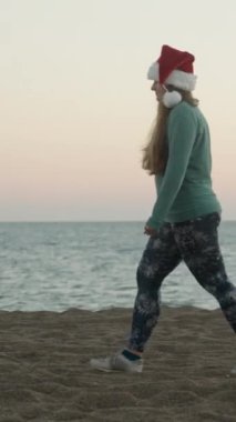 Dikey video. Kırmızı şapkalı genç bir kadın denizin arka planına karşı plaj boyunca yürüyor. Alacakaranlık, günbatımı. Sıcak bir iklimde Noel.
