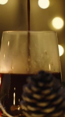 Dikey video. Işıklardan oluşan bir çelengin arkasına sıcak çikolata döküyorum. Mumlar ve dekorasyonlarla dolu bir Noel masası. Yavaş çekim.