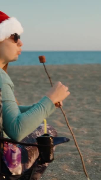 Függőleges Videó Egy Fiatal Télapósapkában Sült Mályvacukrot Eszik Óceán Partján — Stock videók