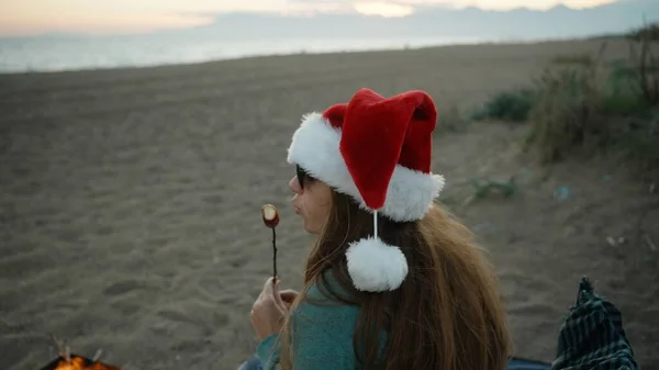 一个戴着圣诞老人红帽子的年轻女子在海滩上吃着炸棉花糖 还在上面吹风 — 图库照片