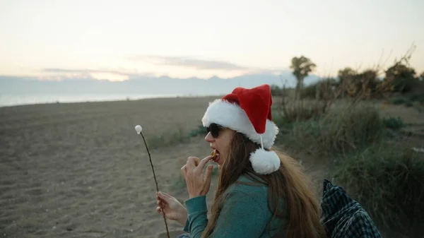 サンタの赤い帽子のビーチにいる若い女性は 揚げたマシュマロを食べて それを吹き飛ばしています — ストック写真