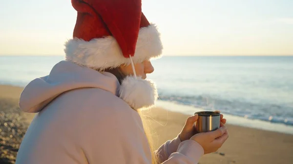 一个凉爽但阳光明媚的圣诞节早晨在海边 一个戴着圣诞礼帽的年轻女子手里拿着一杯热茶保暖 — 图库照片