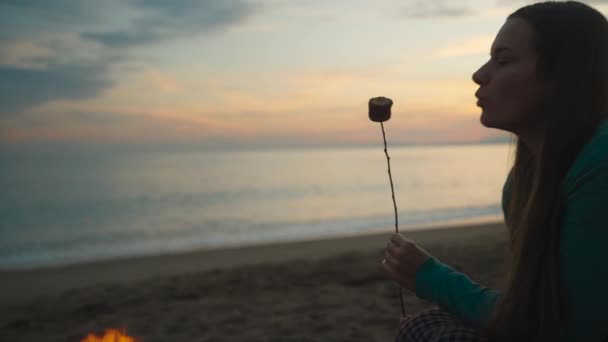 一个年轻的女人在日落时在海边烤棉花糖 她吹在上面 然后继续煎 美丽的日落 — 图库视频影像