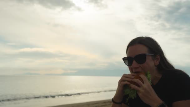 Молода Жінка Окулярах Кусає Бургер Березі Моря Заході Сонця — стокове відео