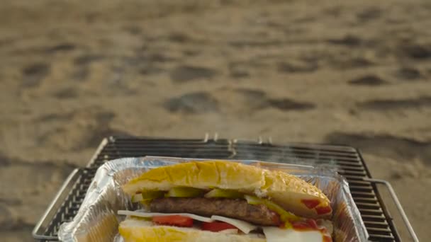 アルミプレートのハンバーガーがグリルに加熱されます 海の背景に対するビーチでのピクニック — ストック動画