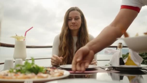 ウェイターは若い女性の皿を食べ物を持って来て 彼女は彼女の手をこすります 海沿いのオープンエアレストラン — ストック動画