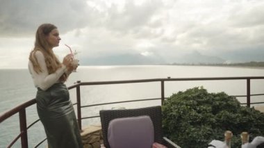 Genç kadın bir kokteyl içiyor. Deniz ve şehir, yüksek bir dağdan çok uzakta..