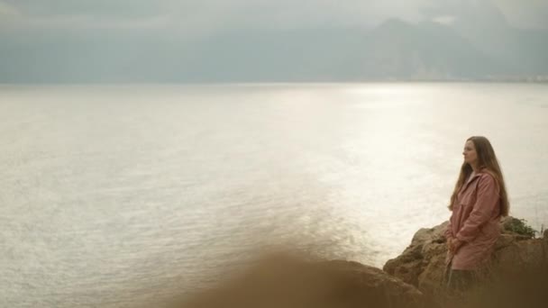 ピンクのレインコートに乗った若い女性が 海の崖の端に座り 背景にある山々を眺めていました 太陽の光が水の上に — ストック動画