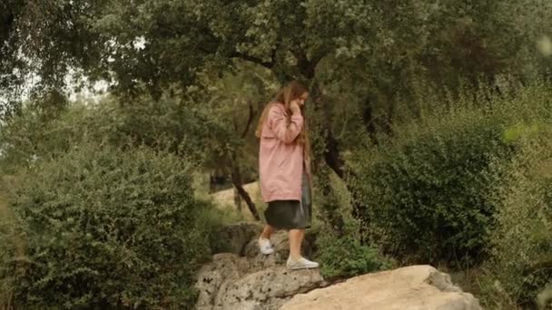 一位身穿粉色雨衣和皮裙的年轻女子走在公园里 她走在岩石上 — 图库视频影像