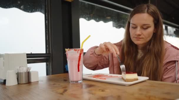 若い女性がカフェでジャケットに座ってチーズケーキを食べている テーブルの上にイチゴミルクシェークがあります — ストック動画