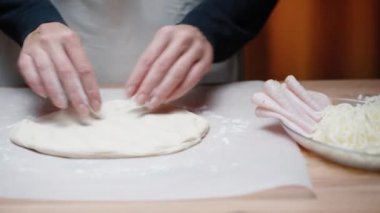 Genç kadın pizza pişiriyor, ellerini masaya koyup hamuru yuvarlıyor, yanında bir dolgu, jambon ve peynir var..