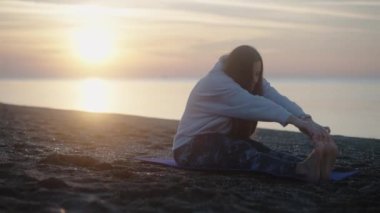 Gün doğumunda genç bir kadın, deniz kıyısındaki bir yoga minderinde geriniyor. Güneş batıyor ve arkasında.
