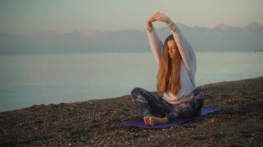 Deniz kıyısında şafak vakti, mor yoga minderindeki genç bir kadın güzel dağların arkasında nişanlanıyor.
