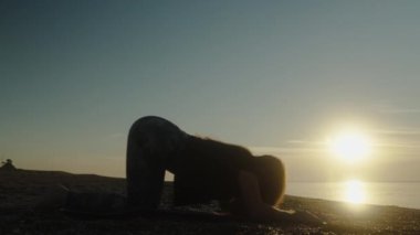 Şafakta deniz kenarında yoga. Genç kadın bir köpek pozisyonunda eğiliyor. Güneşin karşısındaki siluet.