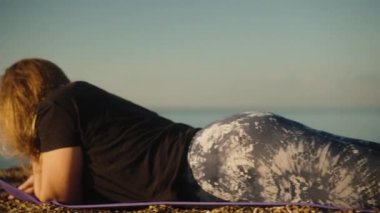 Spor taytlı genç bir kadın deniz kıyısında bir paspasın üzerinde yatıyor, bacakları kalkık..