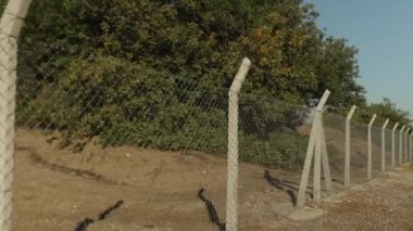Güneşli bir günde tel örgü ve dikenli tellerden yapılmış bir çit boyunca sürüyorum. Arkasında yasak bölge, çöl ve ağaçlar var.